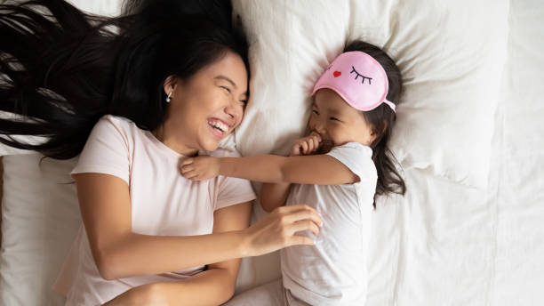 Lying in bed asian mother play with little daughter Above top view cheerful Asian mother tickling play with little toddler daughter in pink eye sleeping mask on head, happy family lying in bed awakened after night sleep feels refreshed and vivacious hot vietnamese women pictures stock pictures, royalty-free photos & images