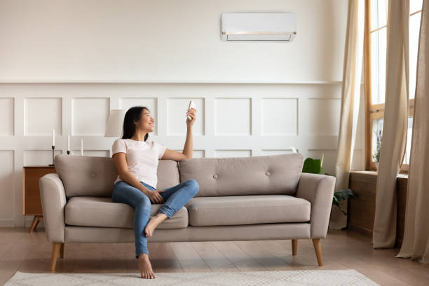 istock Asian woman switching on air conditioner while resting seated indoors 1220837357