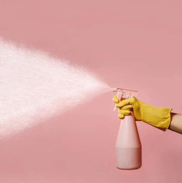 A hand in a silicone household glove holds a liquid spray bottle on a pink background with space for text. A stream of water hits from the atomizer. The concept of maintaining cleanliness and order in the house.