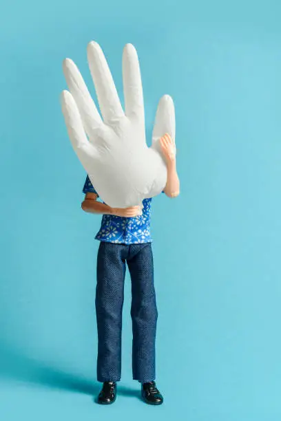 A doll figure holds a puffed-up medical glove while standing against a blue background.