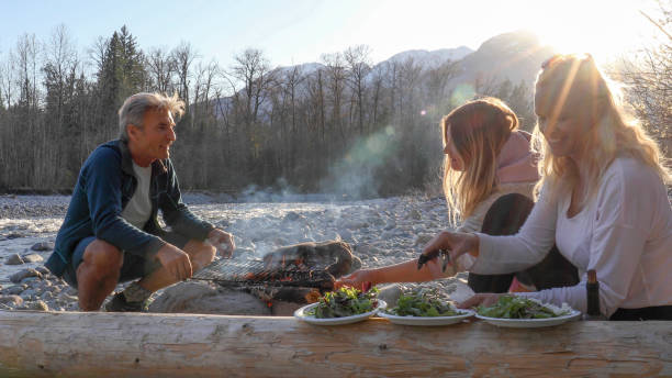 family has picnic over bonfire on river bend - bonfire beach fire barbecue imagens e fotografias de stock