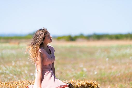 Countryside landscape farm field in Tuscany, Italy with young girl woman happy sitting on hay bale in dress with hair in wind