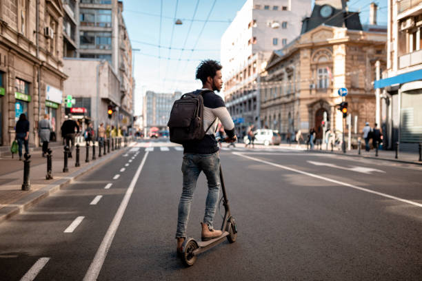 junger mann mit lockigem haar fährt einen elektrischen push-scooter durch die stadt - tretroller stock-fotos und bilder