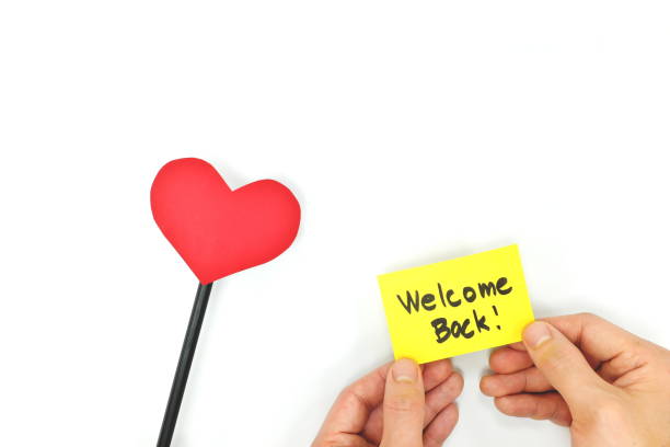 Hand holding a welcome back note on yellow card with a red heart. Back to work, office, business and school. White background.
