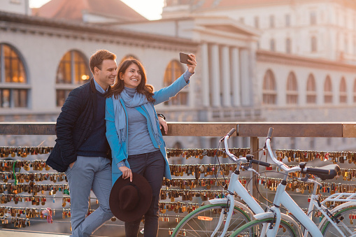 Young smiling couple in love standing on bridge and taking a selfie with cell phone.