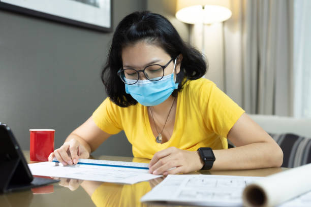 istock Asian female Architect wear eyeglasses using scale ruler for measurement on her blueprints during quarantine in pandemic Coronavirus. Young Woman interior designer in yellow shirt, blue surgical mask working from home. 1220886850