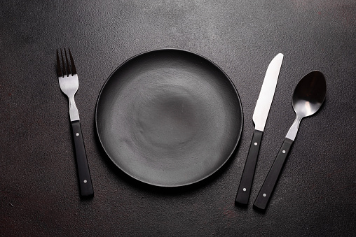 Set of tableware ready for the meal with black copy space. Metal knife, fork, spoon and plate on the dark background