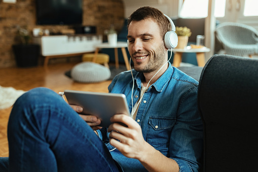 Happy man relaxing at home while using touchpad and listening music over headphones.