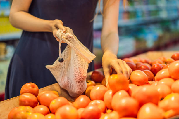 a woman chooses tomatoes in a supermarket without using a plastic bag. reusable bag for buying vegetables. zero waste concept - tomato women green market imagens e fotografias de stock