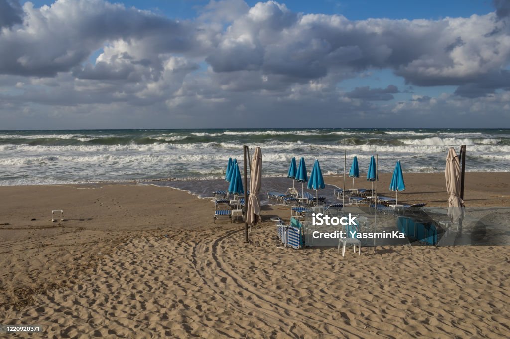 Empty beach in the autumn, Crete, Greece Sandy beach with a group of deckchairs and sun umbrellas with no people in the autumn, during a rainy and stormy day. Waves of the sea and intense colorful clouds. Heraklion, Crete, Greece. Greece Stock Photo