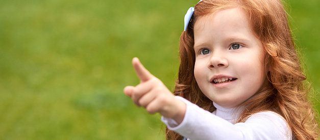 Beauty little woman portrait outdoor. Green background with copyspace. Girl side point. Red hair toodler. Elegance poster kid.