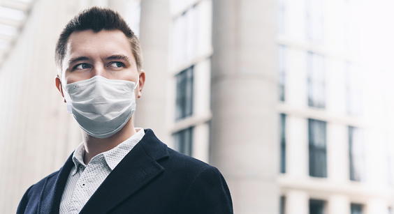 Businessman and coronavirus epidemic. Close up portrait of young business man in a disposable facial mask. A man defends himself against covid 19 on the big city street. Modern buildings at background
