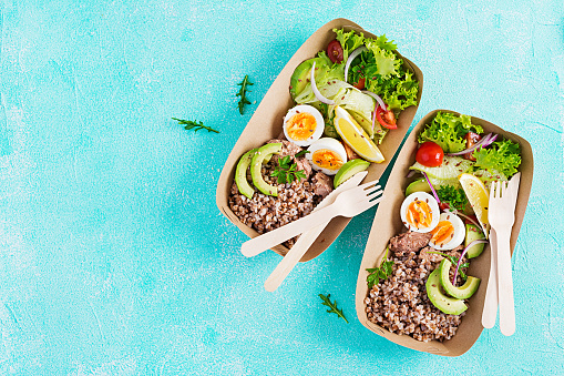 Healthy food delivery. Lunch- Buckwheat porridge, sliced tuna, boiled egg and fresh vegetable salad in zero waste containers  on blue background. Top view, flat lay