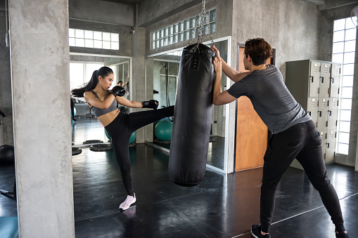 Asian Female boxer punch kick sand bag held by personal trainer at fitness gym. Athletic woman practice Thai boxing with coach for bodybuilding and healthy lifestyle concept.