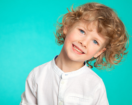 Portrait of a beautiful curly-haired boy with a European appearance on a background of sea-green. The child tilts his head and smiles and laughs. Children's emotions of joy.