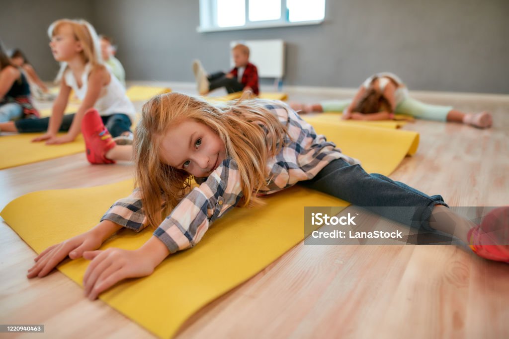 Close Up Portrait Of A Little Cute Girl Sitting On Yoga Mat Stretching Her  Body And Smiling At Camera Group Of Children Doing Gymnastic Exercises In  The Dance Studio Stock Photo 