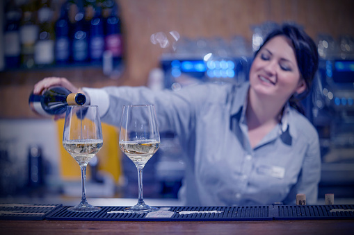 Bartender woman smiling, pours white wine into a glass from a bottle. Shelves with bottles of alcohol in the background.