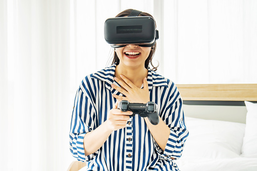 Asia woman wearing VR glasses and sitting in bed in the bedroom. Holding joy stick and playing game by using VR glasses, quarantine due coronavirus pandemic. stay at home.