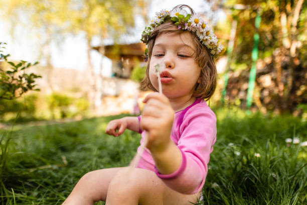 Cheerful little girl sitting on a potty in her backyard and blowing dandelions