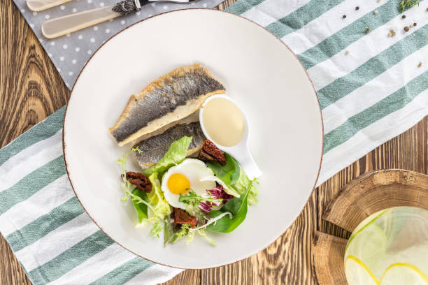 istock Fried white fish fillet with fried egg and greens on white plate on wooden table 1220969998