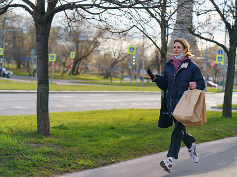 Moscow, Russia - April 23,2020: Young pretty smiling woman wore medical mask and running outdoors. She holding smartphone. Frontal view. Coronavirus Pandemic lifestyle is moving towards completion