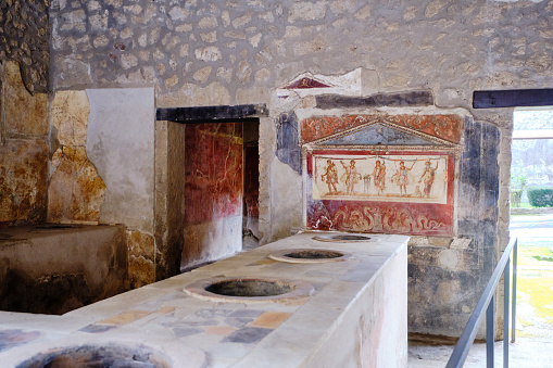 Ancient Thermopolium with Roman Fresco, it is House and Thermopolium of Vetutius Placidus, The thermopolium of Vetutius Placidus opens on via dell’Abbondanza and represents social mobility in Pompeii in Roman times, where merchants and craftsmen also held a high social status, reserved only to landowners in older times. Drinks and hot food were served in this place, as the name indicates, stored in large jars placed in the richly decorated masonry counter of the tavern. photo taken in a street in Pompei called \