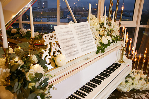 White piano in a restaurant during a wedding celebration. A piano decorated with beautiful bouquets of white flowers and candles in candlesticks.