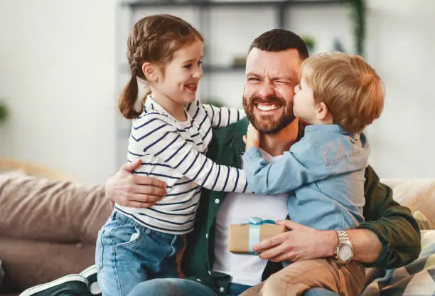 Joyful man with gift box in hand sitting on sofa at home and hugging cheerful son and daughter giving congratulations on fathers day