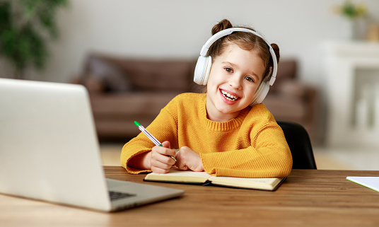 Happy little girl in headphones smiling and making notes in notepad while communicating with teacher through video chat app during online education at home