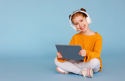 Full length delighted little girl in casual clothes and headphones smiling for camera and listening to music while sitting crossed legged and using tablet on blue background