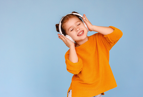 Adorable little girl in casual clothes pressing headphones to ears and smiling while listening to good music with closed eyes against blue background