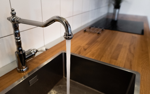Wasting water by leaving a chrome faucet tap running. Water flowing out of a kitchen stainless steel tap into the sink. Water misuse in domestic duties and activities. Overusing household water.
