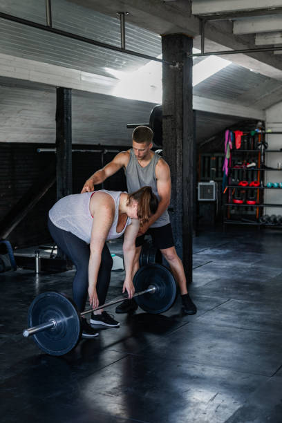 treinamento de força para perda de peso: mulher acima do peso ouvindo seu instrutor de fitness enquanto se exercita - crouching barbell weightlifting weight training - fotografias e filmes do acervo