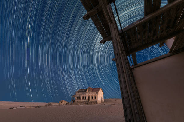 A star trail photograph with an abandoned house in the ghost town of Kolmanskop A beautiful night sky photograph with circular star trails against a deep blue sky, with an abandoned house and desert sand in the background, taken in the ghost town of Kolmanskop, Namibia. kolmanskop namibia stock pictures, royalty-free photos & images