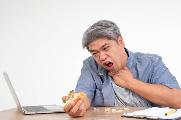 asian man working and eating a burger on office desk and holding his neck after choking foods. concept of a busy businessman cannot work-left balance and not taking care of health eat only junk food - eating sandwich emotional stress food imagens e fotografias de stock