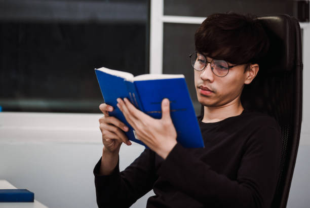 istock Young handsome asian man reading book at work desk late at night, Knowledge and learning concept 1221000199