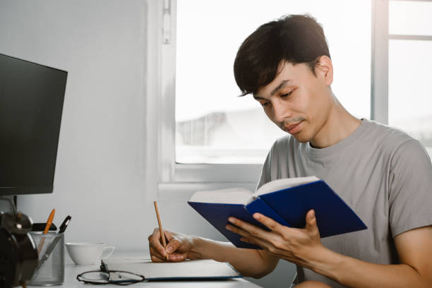 istock Young handsome asian man reading book and writing in note at work desk in free time from working at home, Knowledge and learning concept 1221000263