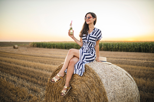 Young happy woman sitting on hay bale and drinking lemonade