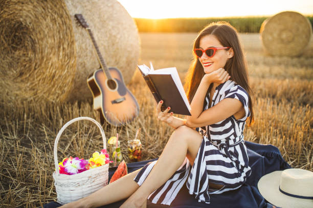 Summer village picnic at sunset. Young woman sitting on blanket, reading a book and having picnic outdoor at agriculture field