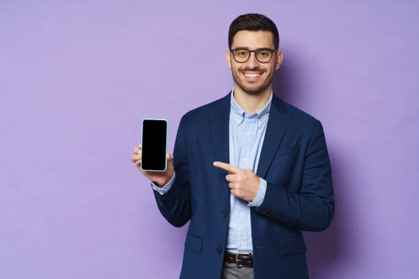 młody biznesmen w garniturze, ubrany w okulary, trzymający pusty ekran smartfona i wskazujący na niego, kopiuj miejsce na reklamę, odizolowany na fioletowym tle - men suit holding human finger zdjęcia i obrazy z banku zdjęć