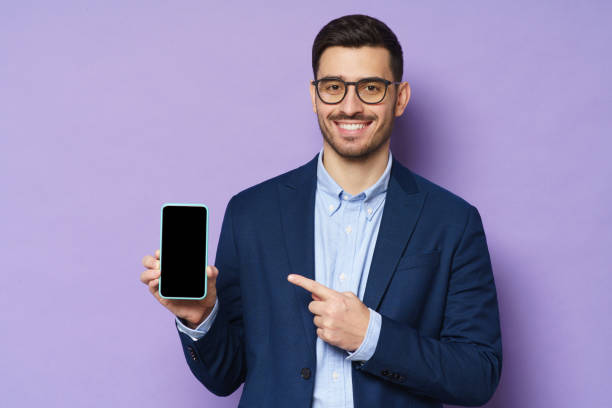 istock Young businessman smiling to viewer, presenting blank screen cellphone, promoting new app on copy space, looking confident, isolated on purple background 1221008861