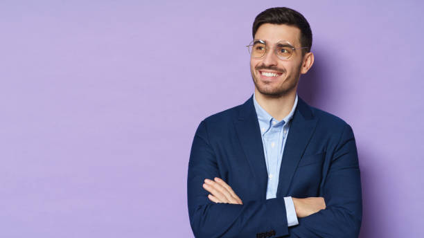 istock Business guy in eyeglasses dressed in blue formal jacket, smiling happily, standing with arms crossed, looking aside, isolated on purple background, copyspace on left 1221008953