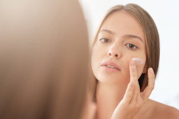 colpo alla testa della giovane bella donna che applica la crema sul viso, guardando il suo riflesso allo specchio, prendendosi cura di se stessa, idratando la pelle al mattino - reflection women contemplation ethnic foto e immagini stock