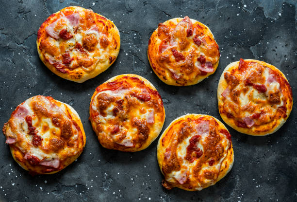 Mini ham, tomato sauce, mozzarella, sun dried tomatoes pizza on a dark background, top view. Delicious breakfast, snack, tapas