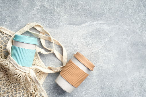 Flat lay eco-friendly takeaway coffee mugs with mesh bag on stone background. Top view bamboo coffee cups and with cotton bag. Zero waste, plastic free, sustainable lifestyle concept