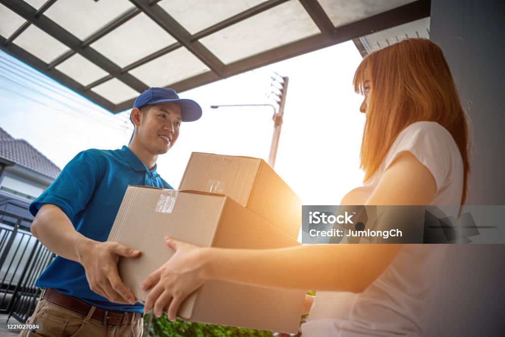 A young Asian deliveryman is delivering a package to a female customer in front of her house. For her to sign to receive the product After she ordered the goods online during the outbreak of the coronavirus or Covid-19 virus. Adult Stock Photo