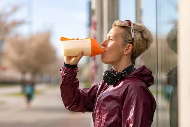 A woman in sports clothing drinking a protein shake. She is wearing a pair of headphones around her neck and a pair of sunglasses on her head.