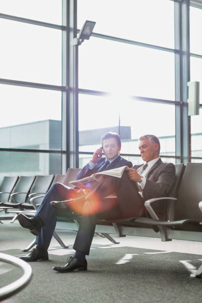 Mature businessman reading news paper while the other businessman is talking on smartphone in airport Mature businessman reading news paper while the other businessman is talking on smartphone in airport newspaper airport reading business person stock pictures, royalty-free photos & images