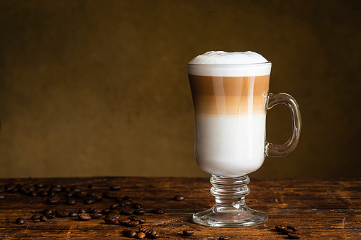 Cafe latte macchiato layered coffee in a see through glass irish coffee glass. The cup is on a wooden table with coffee beans on the table next to the glass.