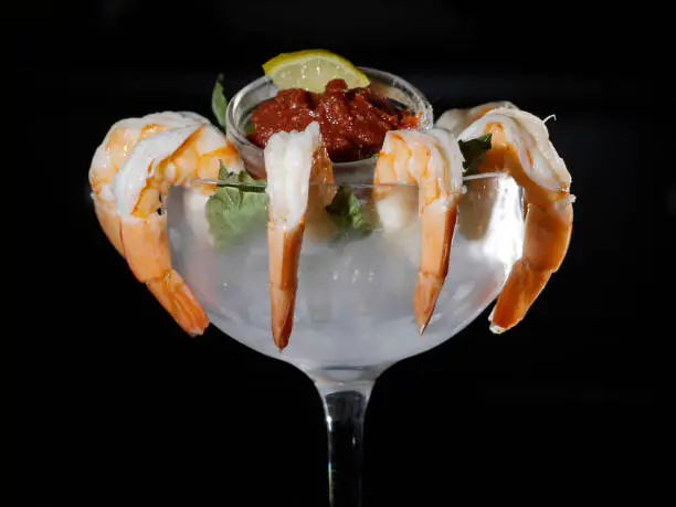 Jumbo Gulf Shrimp cocktail served with cocktail sauce and a yellow lemon wedge in a cocktail-glass filled with ice against a black background.